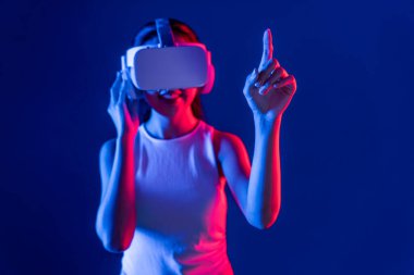 Siberpunk neon ışıklı cihazlarla çevrili zeki bir kadın metaevrene bağlı VR kulaklık takıyor, gelecekçi siber uzay teknolojisi. Sanal objeyi işaret eden bir kadın. Halüsinasyon.