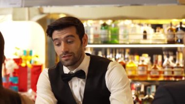 Güzel görünümlü barmen, içki tezgahındaki çiftle samimi bir gülümseme sohbeti yaparken aynı zamanda hoş geldin içkisi ve özel menü listesi öneren çekici profesyonellik. Vinosity.