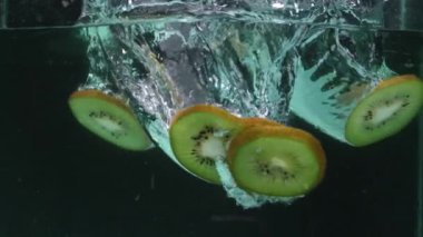 Bir bardak berrak soğuk suya düşen kivi meyvesi dilimlerini kapat. Kivi dilimleri yeşil, siyah tohumlar ve ince beyaz kabuklu. Bazı dilimler hafifçe kıvrılmış. Pabulum.