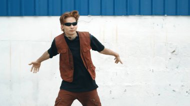 Beyaz arka planda break dans pratiği yapan yakışıklı bir sokak dansçısı. Hip hop tarzı giyinen sporcu, mavi arka planlı bir sokakta müzik eşliğinde ayak sesleri çıkarıyor. Açık hava sporu 2024 hiphop..