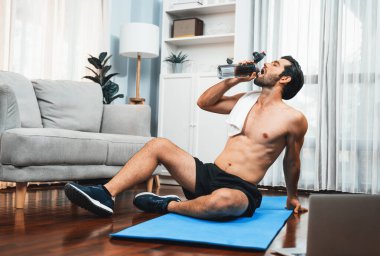 Atletik ve sporcu adam spor paspası üzerinde su içiyor. Vücut egzersizlerini bitirdikten sonra evdeki formda ve sağlıklı spor yaşam tarzı için egzersiz yapıyor. Gaiety evde egzersiz eğitimi.