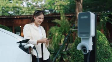 İlkbahar bahçesindeki açık kafede çalışan EV elektrikli araba şarjı, yeşil şehir sürdürülebilirliği ve çevre dostu EV otomobili için genç bir kadınla sürdürülebilir şehir seyahati elektrik ödüyor. Yararlı