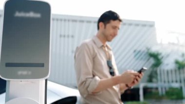 Ağır çekimde çalışan adam, alışveriş merkezinin otoparkındaki kamu EV şarj istasyonunda elektrik için akıllı telefon kullanıyor. EV aracıyla modern çevre ve sürdürülebilir kentsel yaşam tarzı