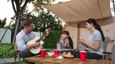 Baba gitar çalarken anne ve kız birlikte kampta oturuyorlar. Ebeveynler, genç nesil çapraz kuşak farklılıklarıyla zaman geçirmek için açık hava kamp aktivitelerini kullanıyorlar. Uyumsuzluk.