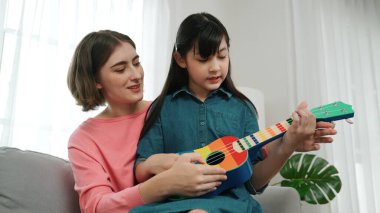 Kafkasyalı bir anne evde akustik müzik öğretirken ukulele çalan mutlu kız. Sevimli çocuk enstrüman öğreniyor. Kafkasyalı mutlu anne ve kız birlikte vakit geçiriyorlar. Pedagoji.