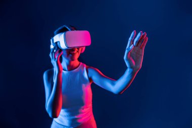 Çevresi siberpunk neon ışıklarıyla çevrili zeki bir kadın metaevrene bağlı VR kulaklık takıyor, gelecekçi siber uzay topluluğu teknolojisi. Sanal objeye el ile dokunan kadın. Halüsinasyon.