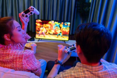 Joystickli birkaç oyuncu dövüş oyunu oynuyor, kadın oyuncu yarışması derecesi arka tarafta TV ekranında modern rahat evinde neon ışıklı oturma odasındaki koltukta oturuyor. İnfobahn.
