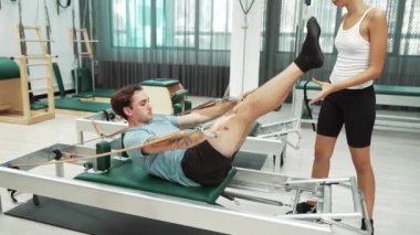 Bir grup müşteri, yenilikçi bir makinede bacak yükseltme işlemi yapıyor. Pilates partneri reformcu pozu veriyor ve egzersiz sırasında dengeyi geliştirmek ve koordinasyonu arttırmak için kayış takıyor. Alışkanlık.