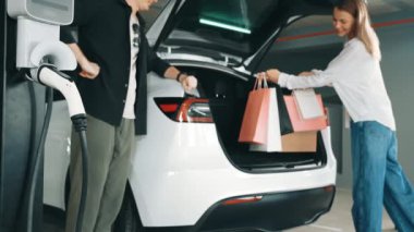 Yavaş çekim çift EV elektrikli arabayla şehir merkezindeki alışveriş merkezine gidiyor. Şehir merkezinde yeşil, temiz, yeniden şarj edilebilir elektrikli araç iç organları tarafından sürdürülebilirlik yaşam biçimi olarak şarj ediliyor.