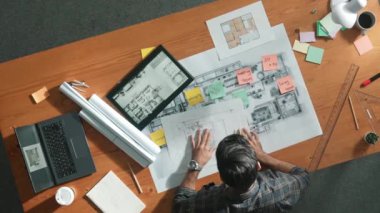 Proje yöneticisinin elinde tabletle toplantı masasında otururken projeye bakışının üst görüntüsü. Çeşitli meslektaşlar toplantıya katılırken, yetenekli mühendisin proje planı üzerinde çalışmasının havadan görüşü. Sempozyum.