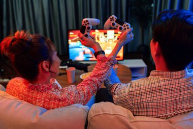 joystickli elleri çapraz oynayan bir çift arka tarafta TV ekranında dövüş oyunu oynuyor, modern rahat evinde neon ışıklı oturma odasında kanepesinde oturuyor. İnfobahn.