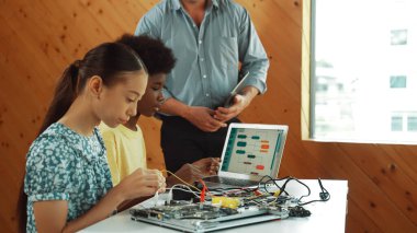 Öğretmen ayakta dururken kodlamayı ve robot yapısını açıkla. Çeşitli gençler ana kartı tamir ediyor ve dizüstü bilgisayarın programlama ekranına bakarken elektronik alet kullanıyorlar. Öğretim.