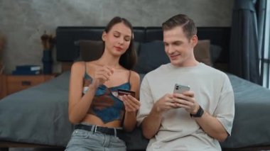 Genç çift, kredi kartıyla ödemek için internet üzerinden ödeme uygulaması ve dijital cüzdan kullanarak yatak odasında oturuyor. Mobil internet üzerinden alışveriş ve modern alımlar. Düzenle