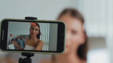 Genç sosyal medyum kadın moda videosu yapıyor. Blogcu kameraya selfie çubuğu ve ışık halkası kullanarak gülümserken, aynı zamanda ikna edici çevrimiçi kıyafetlerin izleyicilere veya takipçilere vlog satmasını sağlıyor. Düzenle