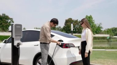 Genç çift, baharda açık yeşil şehir parkındaki şarj istasyonundan elektrikli araba aküsünü şarj ediyor. Enerji sürdürülebilir kentsel seyahat yaşam tarzı için şarj edilebilir EV araba
