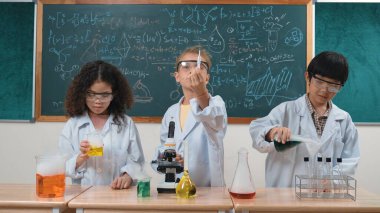 Çocuklar laboratuvarda karatahta başında dururken fen dersinde deney yapıyorlar. Mutlu öğrenci biyokimyasal sıvıyı keşfederken ve incelerken ve karıştırırken. Pedagoji.