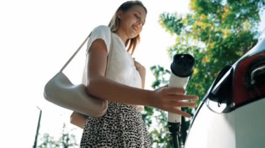 Yavaş çekim güneş ışığı arka plan aydınlatması EV elektrikli arabasıyla gezen kadın yazın yeşil sürdürülebilir şehir bahçesinde şarj oluyor. Elektrikli araç iç organlarından yeşil kentsel sürdürülebilirlik yaşam tarzı