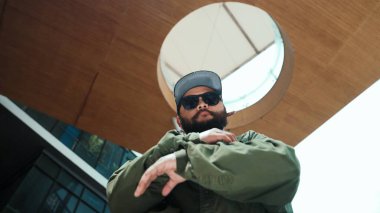 Kameraya bakarken Asyalı yakışıklı hipsterin hip hop pozu. Profesyonel sokak dansçısının alışveriş merkezinde serbest dans edişinin düşük açılı görüntüsü. Açık hava sporu 2024. Hiphop.