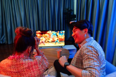 joystickli bir çift, TV ekranında dövüş oyunu oynuyor, erkek, kız arkadaşıyla yarışma seviyesini geri kazanıyor, modern evdeki neon ışıklı oturma odasında kanepeye oturuyor. İnfobahn.