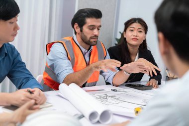 Mimari proje üzerinde birlikte çalışan çeşitli inşaat mühendisi ve müvekkil grupları, inşaat planını gözden geçirip toplantı masasında plan oluşturuyorlar. Tedbirli