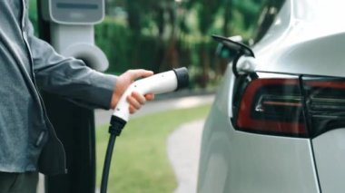 Yavaş çekim adamı yeşil sürdürülebilir şehir parkı yaz bahçesinde EV elektrikli otomobilini kentsel sürdürülebilirlikteki gençlerin yaşam tarzı olarak şarj et.