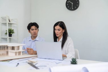 Profesyonel Asyalı erkek mimar ev modelinin uzunluğunu ölçmek için cetvel kullanırken, beyaz tenli genç meslektaşı ev modeliyle toplantı masasındaki verileri analiz etmek için dizüstü bilgisayar kullanıyor. Tertemiz..