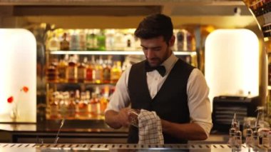 İyi görünümlü, gülümseyen, bardaki lüks içki tezgahında cam silen barmen profili. Bar gece kulübünde sıcak karşılayan profesyonel barmen kostümünü poz vermek için kameraya bakıyor. Vinosity.