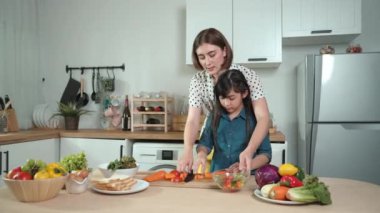 Zeki beyaz anne ve Asyalı kız birlikte yemek pişiriyor ve sebze doğruyor ya da akşam yemeği için salata hazırlıyor. Mutlu anne ve kız taze yemekle sağlıklı yemek yapıyorlar. Sağlıklı gıda konsepti. Pedagoji.