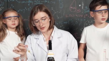 Öğretmen tavsiye verirken beyaz çocuk kimyasal sıvıyı karıştırıyor. Profesyonel eğitmen labaratuar kıyafeti giymiş, renkli solüsyonla dolu deney kabıyla sofrada çeşitli öğrencileri arıyor. Etkinlik.