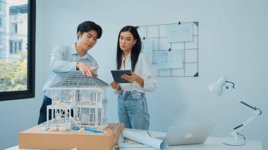 Güzel, genç, beyaz bir proje yöneticisi Asyalı mühendise, mühendislik masasında ev modelini ölçerken tablet kullanarak bina detaylarını anlattı. Tertemiz.