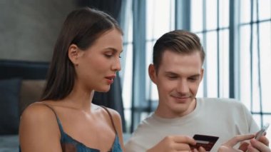 Genç çift, kredi kartıyla ödemek için internet üzerinden ödeme uygulaması ve dijital cüzdan kullanarak yatak odasında oturuyor. Mobil internet üzerinden alışveriş ve modern alımlar. Düzenle