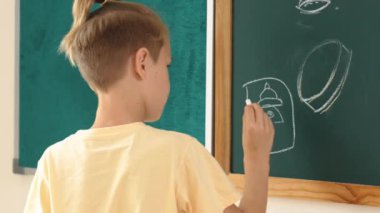 Kafkasyalı enerjik zeki çocuk sınıfta tahtada dururken hayal gücünü ifade etmek ve geliştirmek için resim çiziyor. Sanat dersinde karatahtaya yazı yazan çekici ilkokul öğrencisi. Pedagoji.