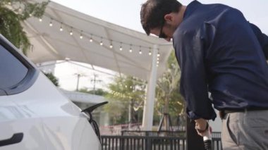 Genç adam, EV şarj aletini şehrin alışveriş merkezinin otoparkındaki şarj istasyonundan elektrik aküsünü şarj etmek için kullandı. Sürdürülebilir şehirde EV aracı tarafından şarj edilebilir enerji olarak kullanıldı.