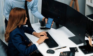Gülümseyen Asyalı işadamı telefondaki dinamik borsa verilerini bilgisayarda kar değeri değerinin analizi için arayan kadın ortağa gösteriyor. Ofisteki ekranlarla karşılaştırınca. İnfobahn.