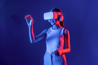 Çevresi siberpunk neon ışıklarıyla çevrili zeki bir kadın metaevrene bağlı VR kulaklık takıyor, gelecekçi siber uzay topluluğu teknolojisi. Sanal objeye el ile dokunan kadın. Halüsinasyon.