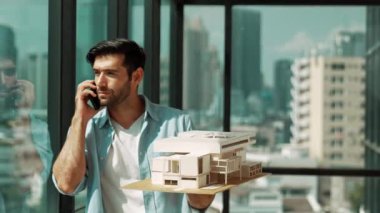 Gülümseyen Kafkasyalı mühendis ev modelini sağa sola çeviriyor ve iş arkadaşına telefon ederken hata noktasını kontrol ediyor. Profesyonel yönetici teftiş, kontrol, mimari modele bakmak. İzleme