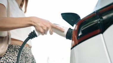 Yavaş çekim güneş ışığı arka plan aydınlatması EV elektrikli arabasıyla gezen kadın yazın yeşil sürdürülebilir şehir bahçesinde şarj oluyor. Elektrikli araç iç organlarından yeşil kentsel sürdürülebilirlik yaşam tarzı