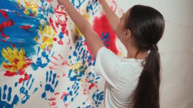 Asyalı kız resim dersinde duvara kırmızı renk damgalamak için elini kullanır. Çok kültürlü bir lise öğrencisi beyaz gömlek giyerken boya odasında renkli el izleri olur. Yaratıcı aktivite konsepti. Öğretim