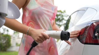 Kapalı alan kadını, doğal yeşil parktaki otoparkta EV elektrikli araba aküsünü şarj ediyor. Sürdürülebilir ve çevre dostu seyahat için şarj edilebilir araç için temiz enerji teknolojisi. Eşzamanlama