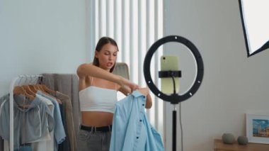 Genç sosyal medyum kadın moda videosu yapıyor. Blogcu kameraya selfie çubuğu ve ışık halkası kullanarak gülümserken, aynı zamanda ikna edici çevrimiçi kıyafetlerin izleyicilere veya takipçilere vlog satmasını sağlıyor. Düzenle