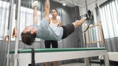 Eğitmen rehberliğinde Pilates Cadillac 'ta ters bacak yükseltme pratiği yapan sağlıklı bir müşteri. Çekirdek dayanıklılığını ve üst vücut dengesini arttırmak için Cadillac 'ı geren adam. Alışkanlık.