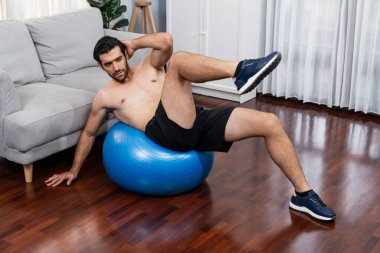 Sporcu ve atletik bir adam evde vücut egzersizi yaparken spor topunun üzerinde oturarak formda fiziki ve sağlıklı spor yaşam tarzına uygun bir yaşam sürüyor. Gaiety ev egzersizi antrenman konsepti.