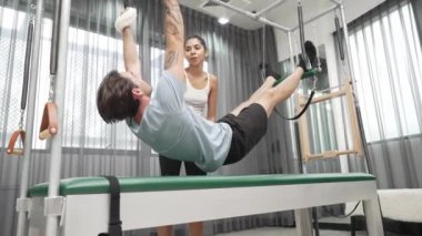 Eğitmen rehberliğinde Pilates Cadillac 'ta ters bacak yükseltme pratiği yapan sağlıklı bir müşteri. Çekirdek dayanıklılığını ve üst vücut dengesini arttırmak için Cadillac 'ı geren adam. Alışkanlık.