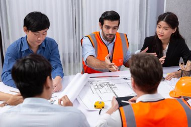 Mimari proje üzerinde birlikte çalışan çeşitli inşaat mühendisi ve müvekkil grupları, inşaat planını gözden geçirip toplantı masasında plan oluşturuyorlar. Tedbirli