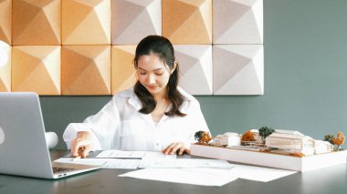 Güzel genç mimar mühendisinin portresi ev modelini ölçerken aynı zamanda dizüstü bilgisayar, proje planı ve masanın üzerine yerleştirilmiş taslağı da alıyor. İş tasarımı konsepti. Yönlendirici.