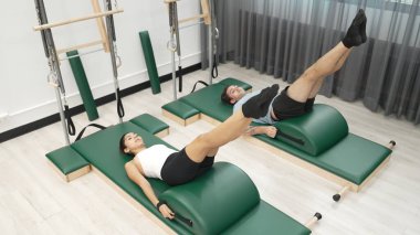 Pilates seansı sırasında iki müşteri yay namlu teçhizatı kullanarak bacak kaldırma pratiği yapıyor. Mutlu antrenör grubu karın kaslarını ve alt karın kaslarını geliştirmek için makas ya da bacak esneme pozu verir. Alışkanlık.