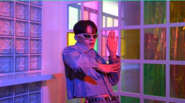 Gece kulübünde kameraya bakarken güneş gözlüklü Asyalı sanatçının serbest dans edişini yakından çek. Mutlu sokak dansçısı dans ediyor ve renkli parlak neon ışıklı ayak izlerini gösteriyor. Mutluluk.