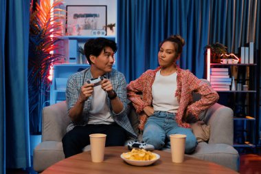 Mutlu çiftin oturduğu kanepe joysticklerle video oyunu oynuyor, meydan okuyan gülümseyen adam, evdeki modern neon ışıklı oturma odasında kazanmak için yumruğunu kaldırıyor. İnfobahn.