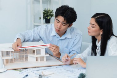 Profesyonel Asyalı erkek mimar ev modelinin uzunluğunu ölçmek için cetvel kullanırken, beyaz tenli genç meslektaşı ev modeliyle toplantı masasındaki verileri analiz etmek için dizüstü bilgisayar kullanıyor. Tertemiz..