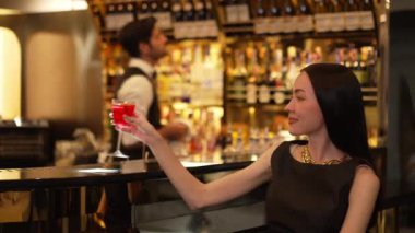 Güzel bir konuk, gece kulübünün mükemmel atmosferinde barmenlerin arka planında çiçeklerle süslenmiş karışık votka Cosmopolitan kırmızı kokteyli ile lüks bir gişede içki içerken kameraya bakıyor. Vinosity.